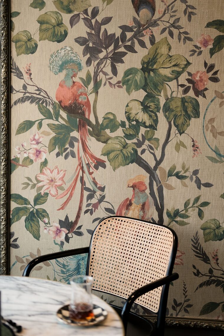 Reading nook with botanical bird-print wallpaper and a cane chair
